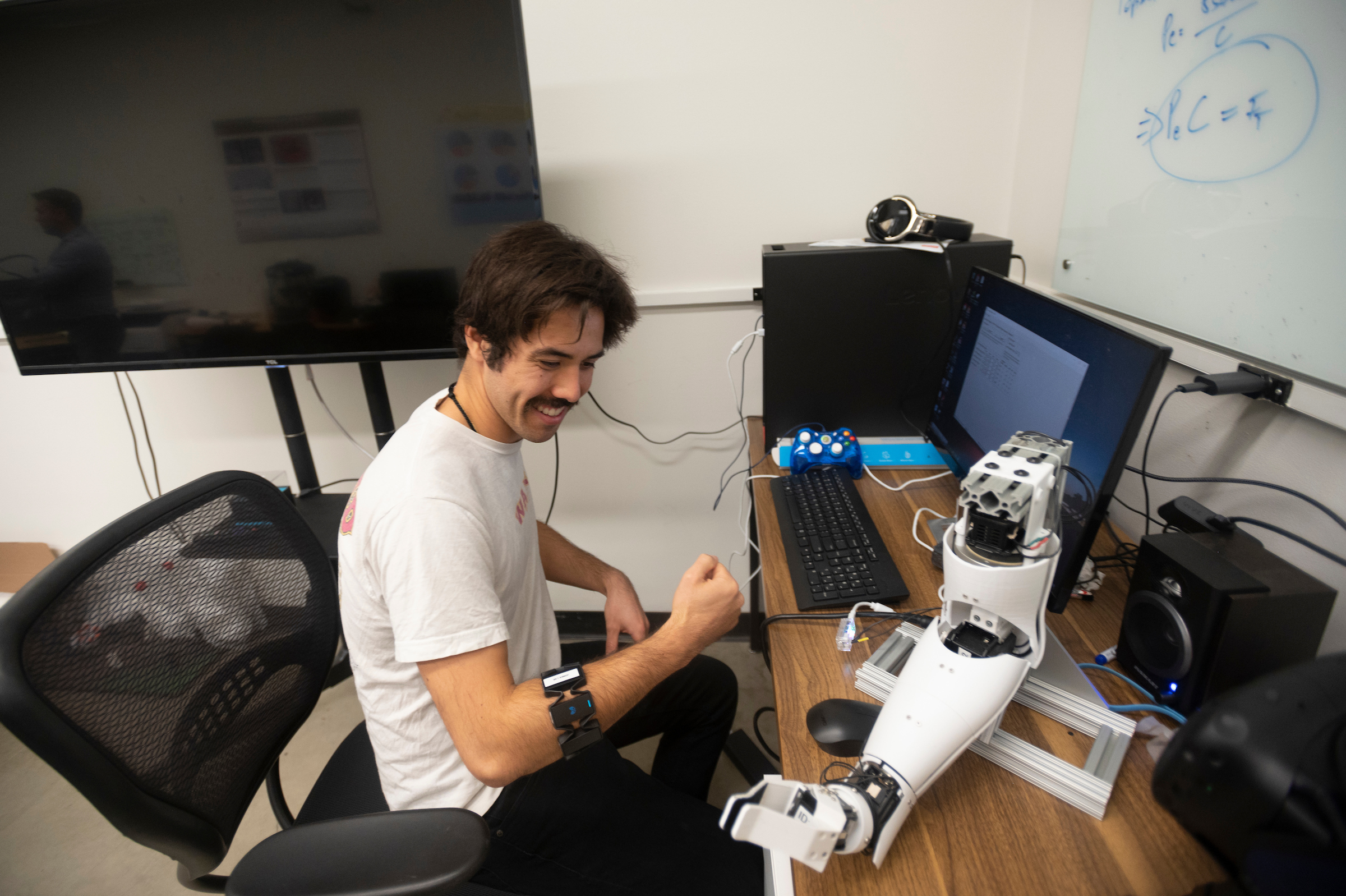 Person sits at a desk wearing a mechanical arm band and looking at a prosthetic arm