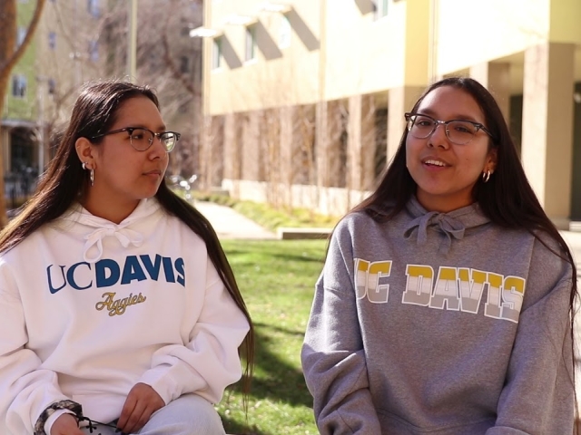 Two twins in UC Davis sweaters sit outside near a building