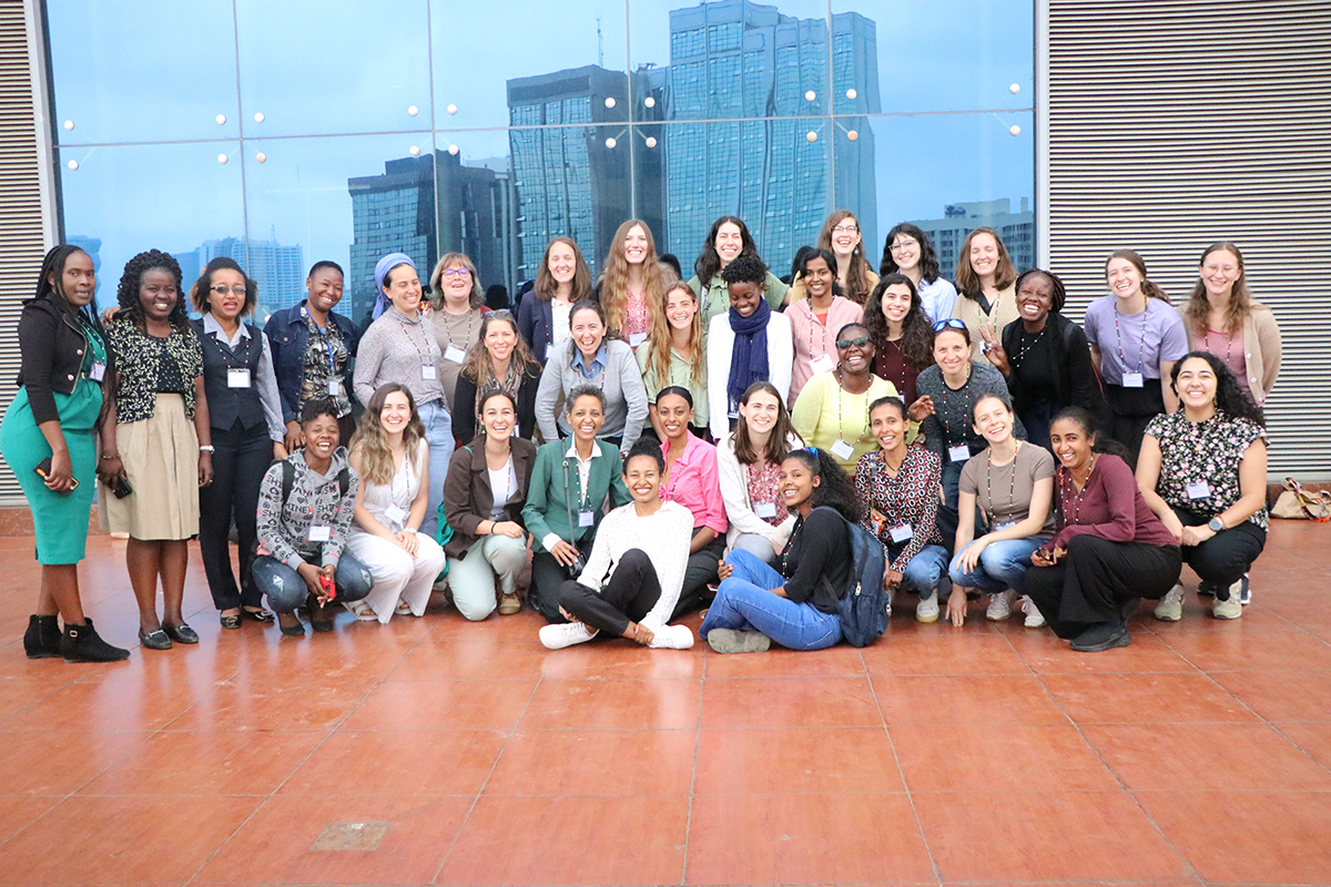 Large group of women pose for a group photo, wearing lanyards and standing by a mirror that shows large buildings reflected