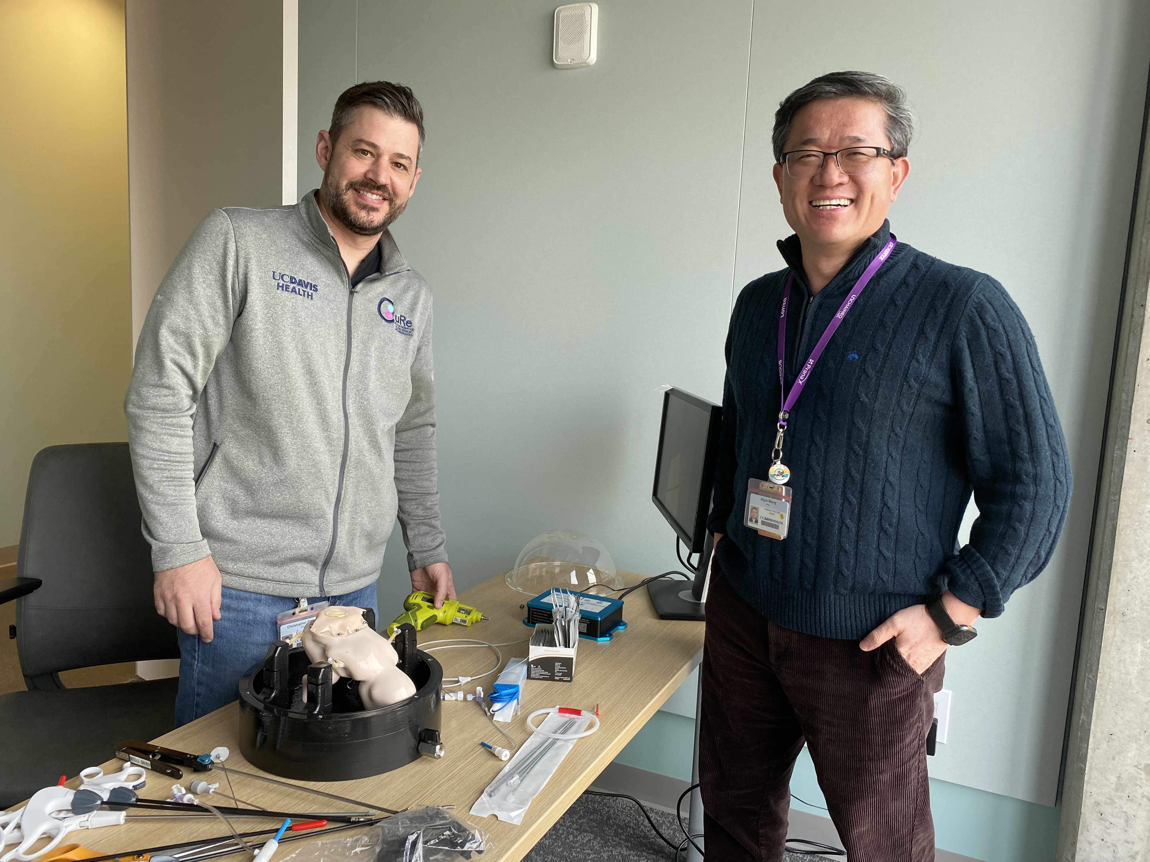 Two men standing by a table with tools, a fetus model, and a computer, smiling at the camera.