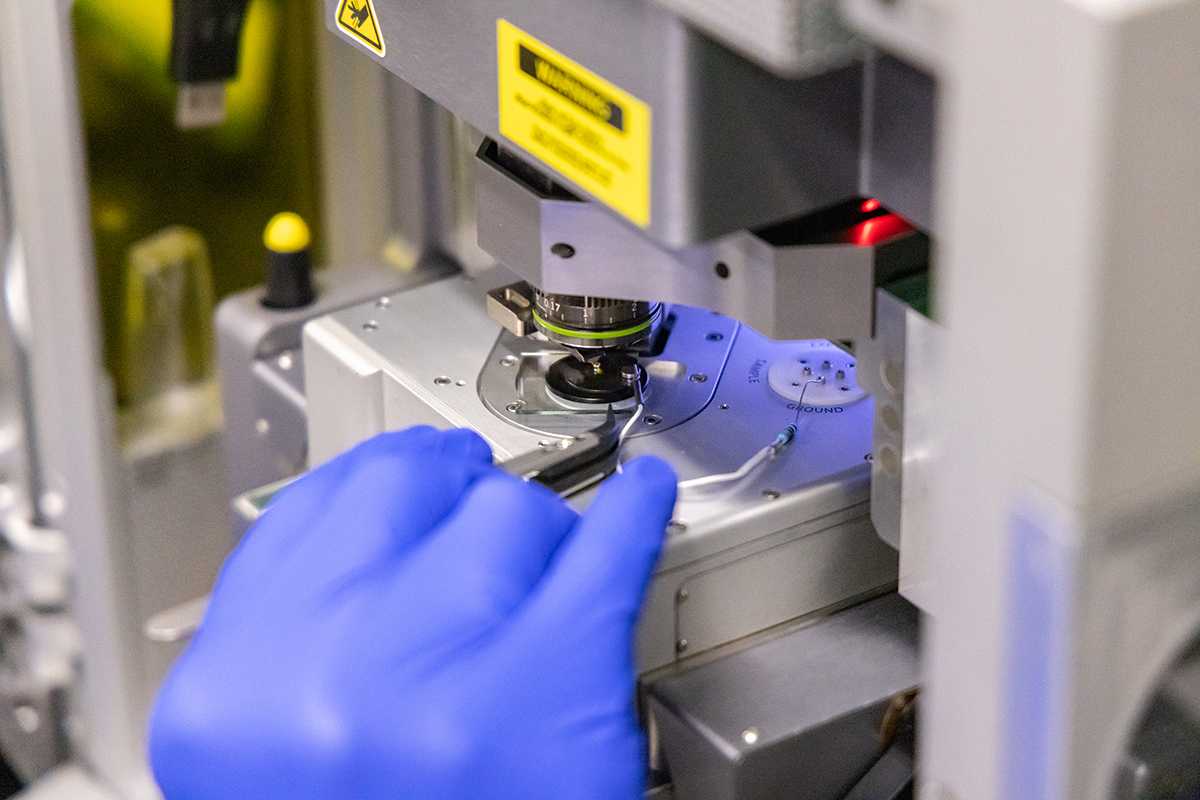 Close up of gloved hand holding tweezers and pushing a glass slide into a microscope