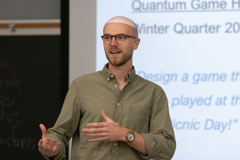 A man gestures while speaking in front of a classroom projector.