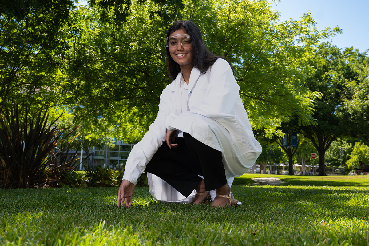 photo of a student wearing a lab coat and PPE crouching down to touch the grass