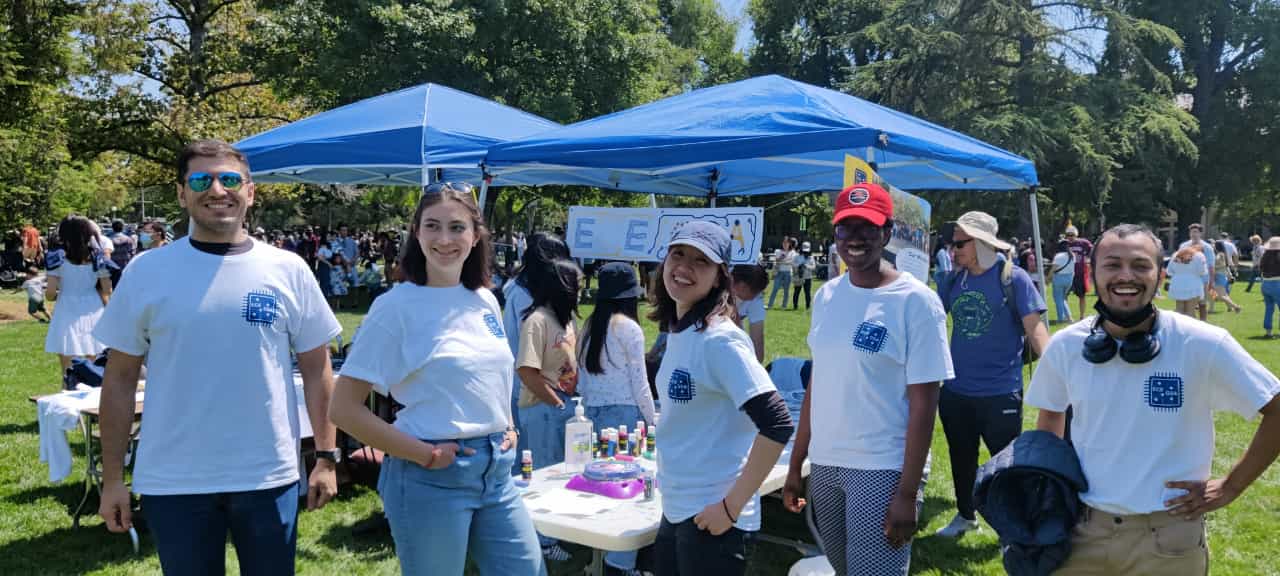 Students in white shirts stand outdoors near blue pop up tents at an event