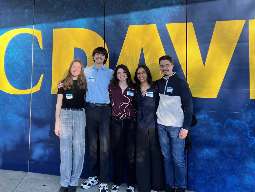 The FirstBite team smiling for a photo in front of a blue wall with large yellow letters that say "UC Davis"