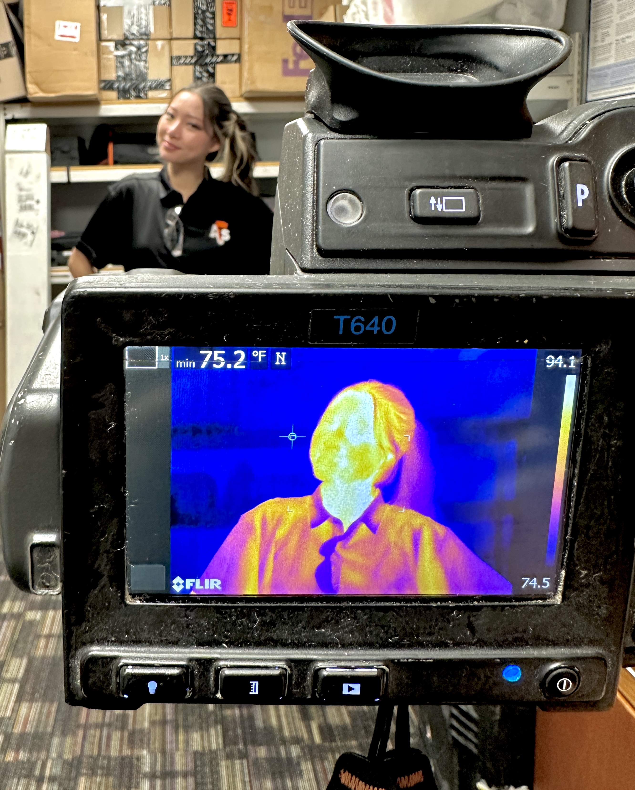 Thermal camera display photograph showing hot-colored face on screen with staff and shelves behind