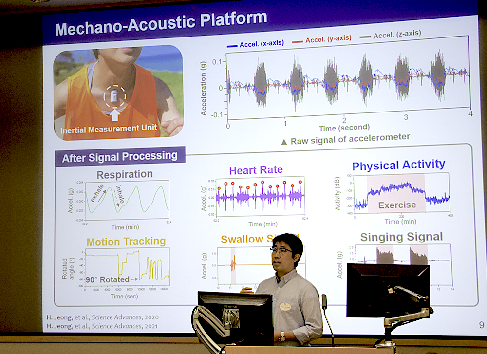 Dr. Jeong giving a presentation in front of a large slide image with graphs for respiration, heart rate, motion tracking, and other metrics. 