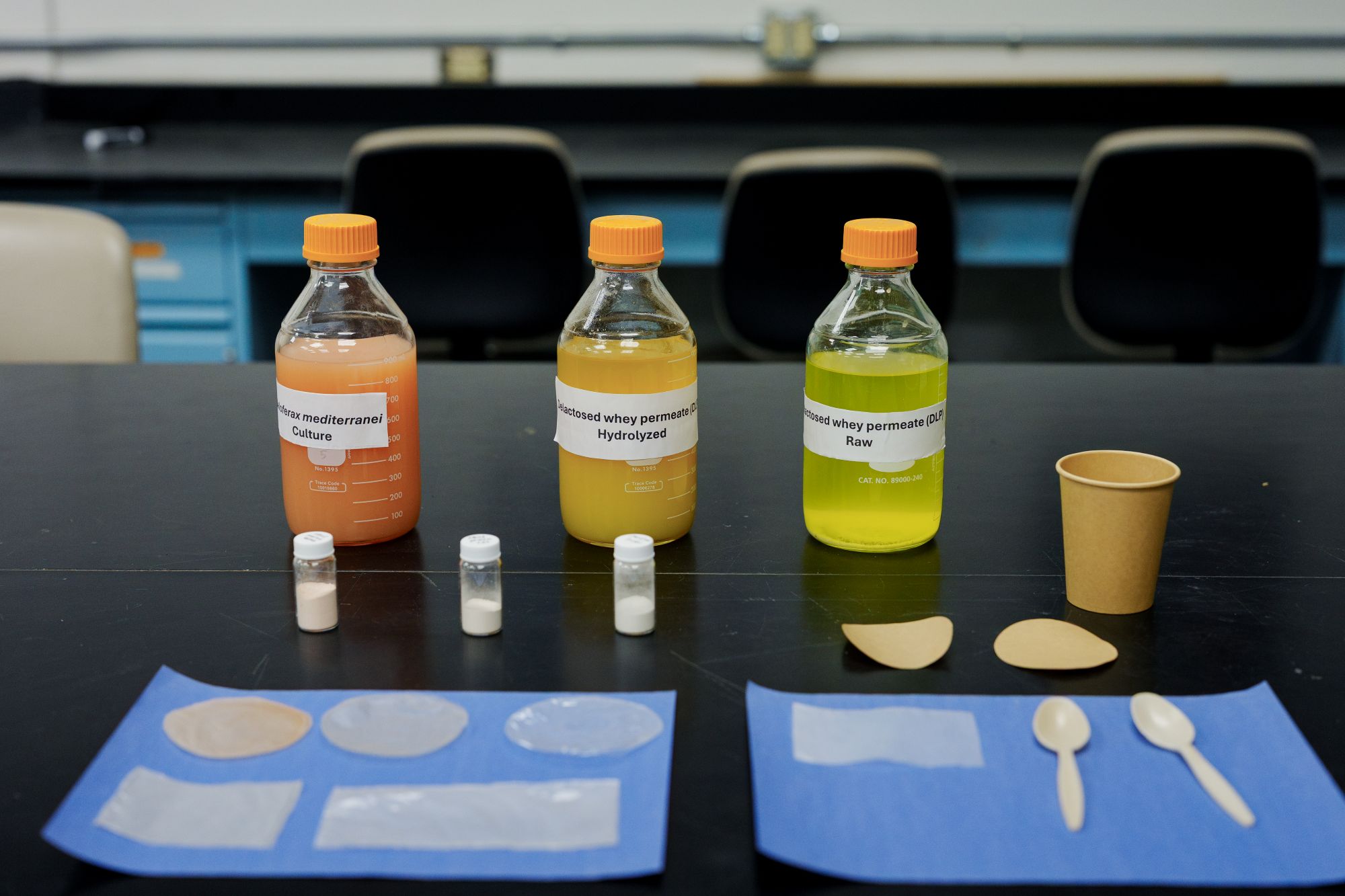 Three labeled bottles, small vials, paper cup and spoons on a black lab table.