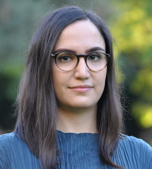 A dark-haired woman with glasses wearing a blue top in front of a natural background