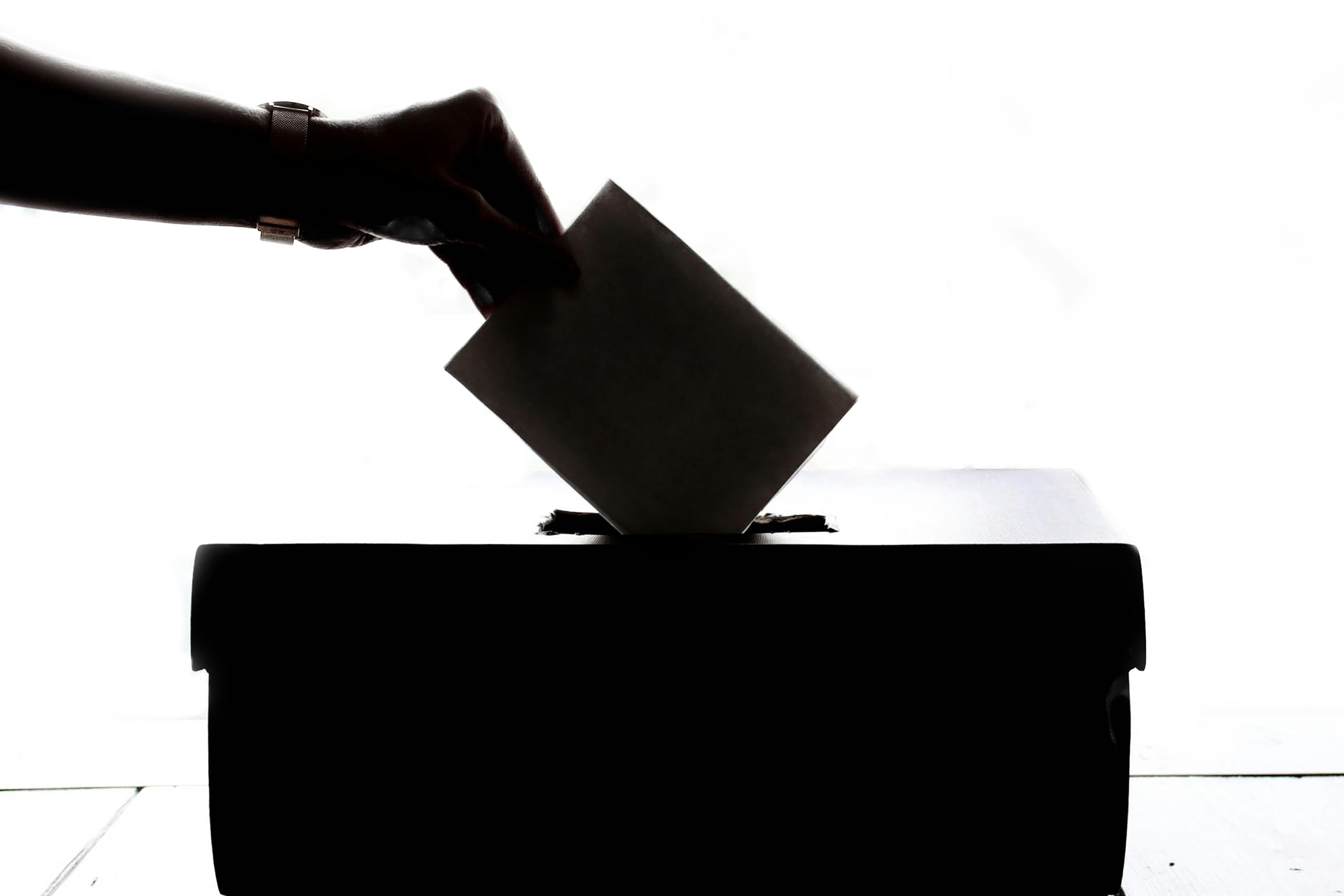 A hand depositing a ballot into a voting box, silhouetted against a bright background.
