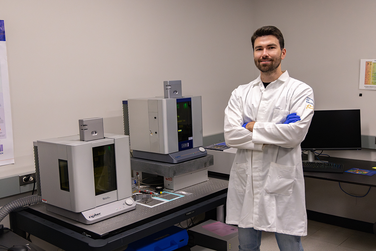 Shelden stands with two atomic force microscopes in the lab, wearing PPE