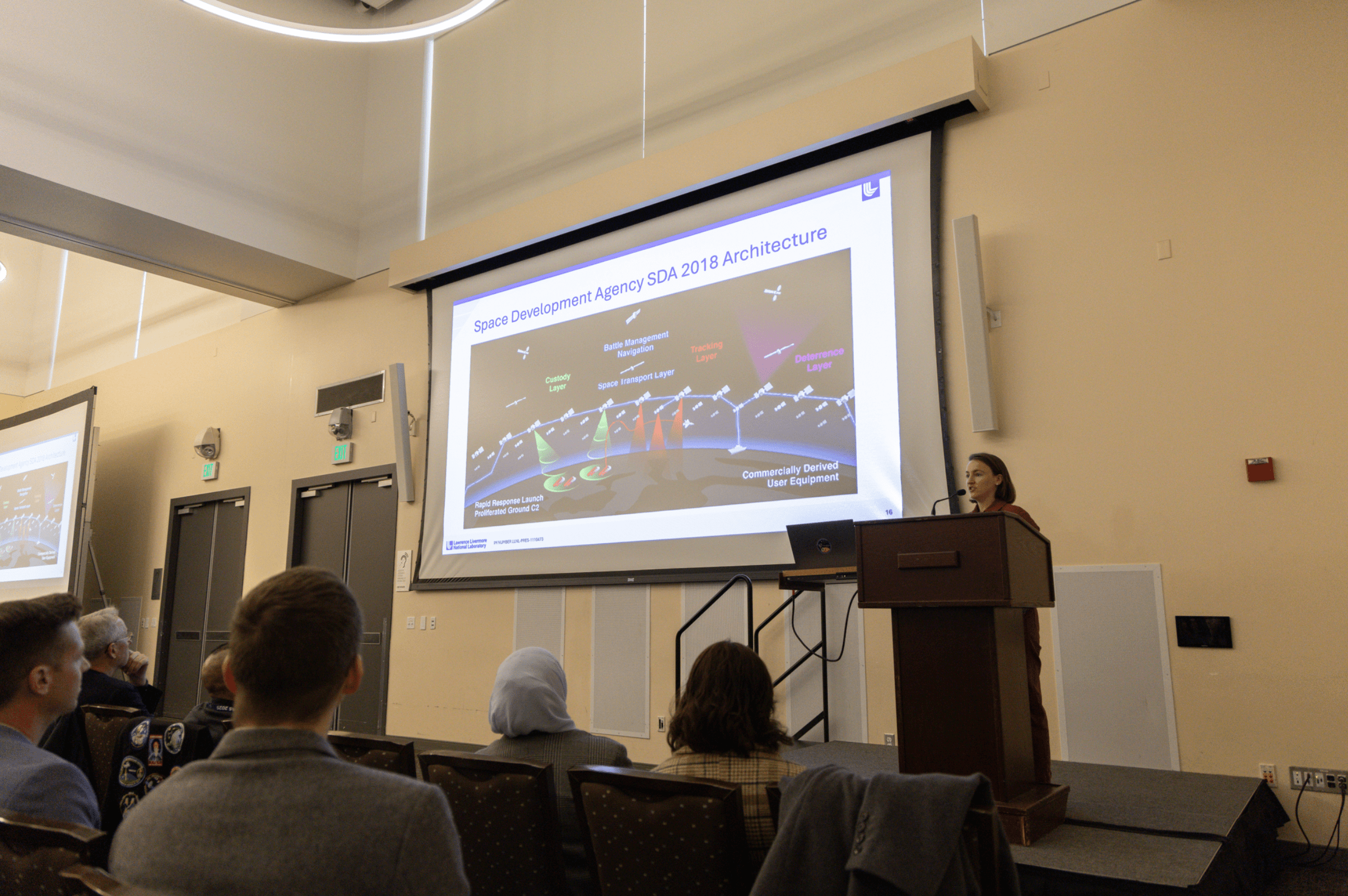 A presenter standing at a podium next to a projector screen showing research