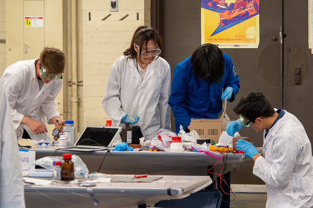 Four students in lab coats, goggles and gloves work in a lab with various materials