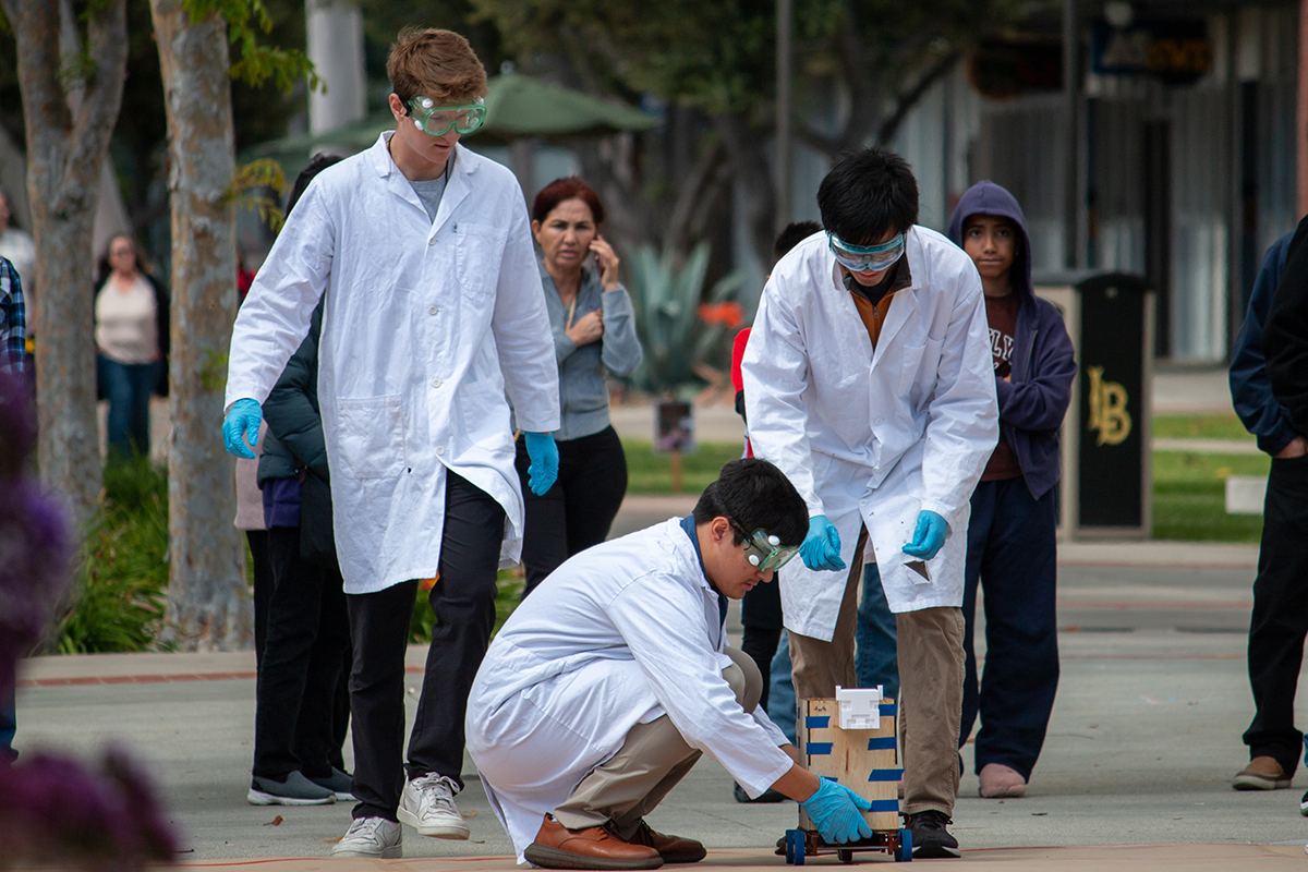 Three students in PPE stand outdoors while one places a car on the ground and people look on in the background