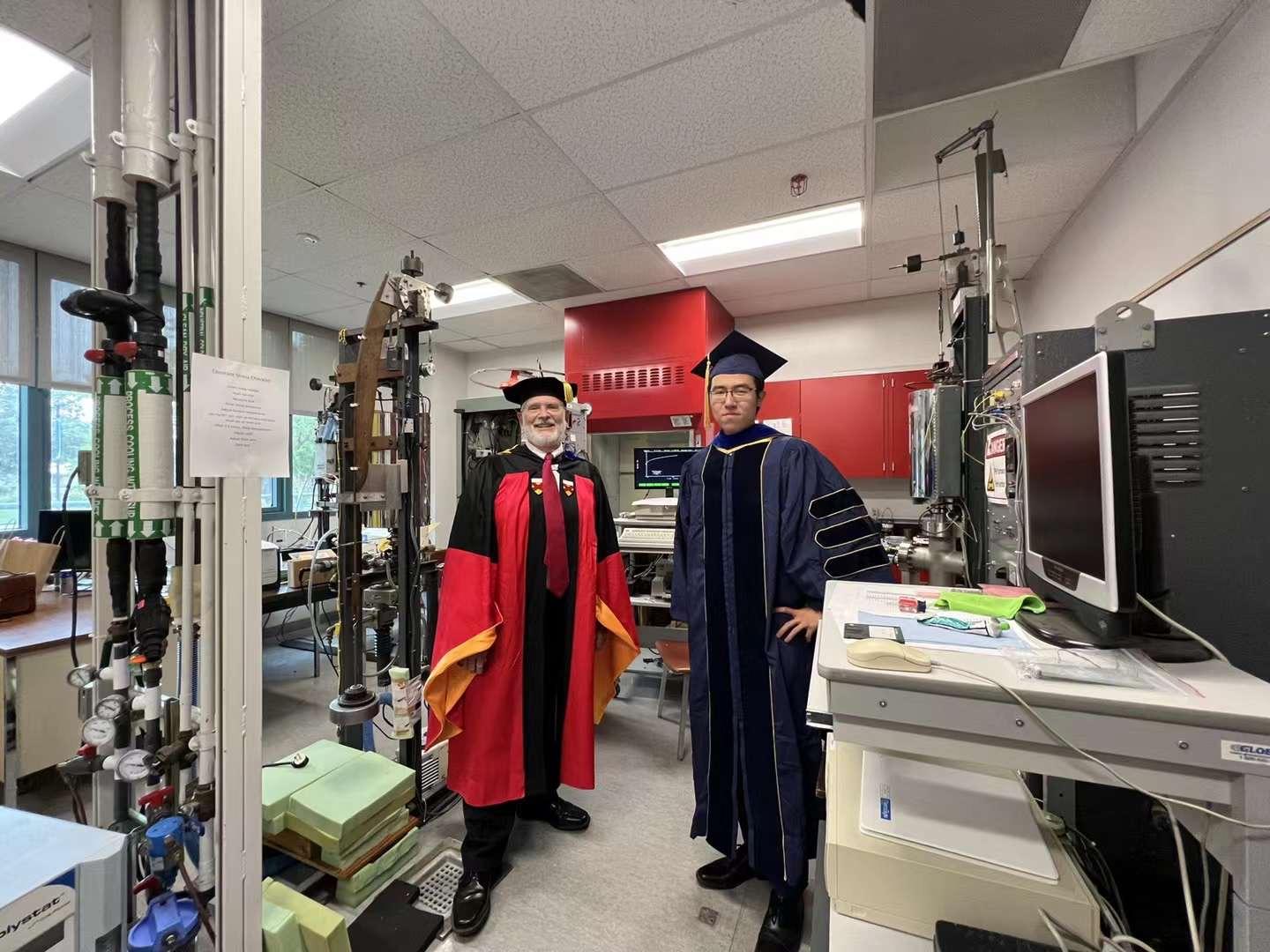 Two people stand in a lab wearing graduation regalia