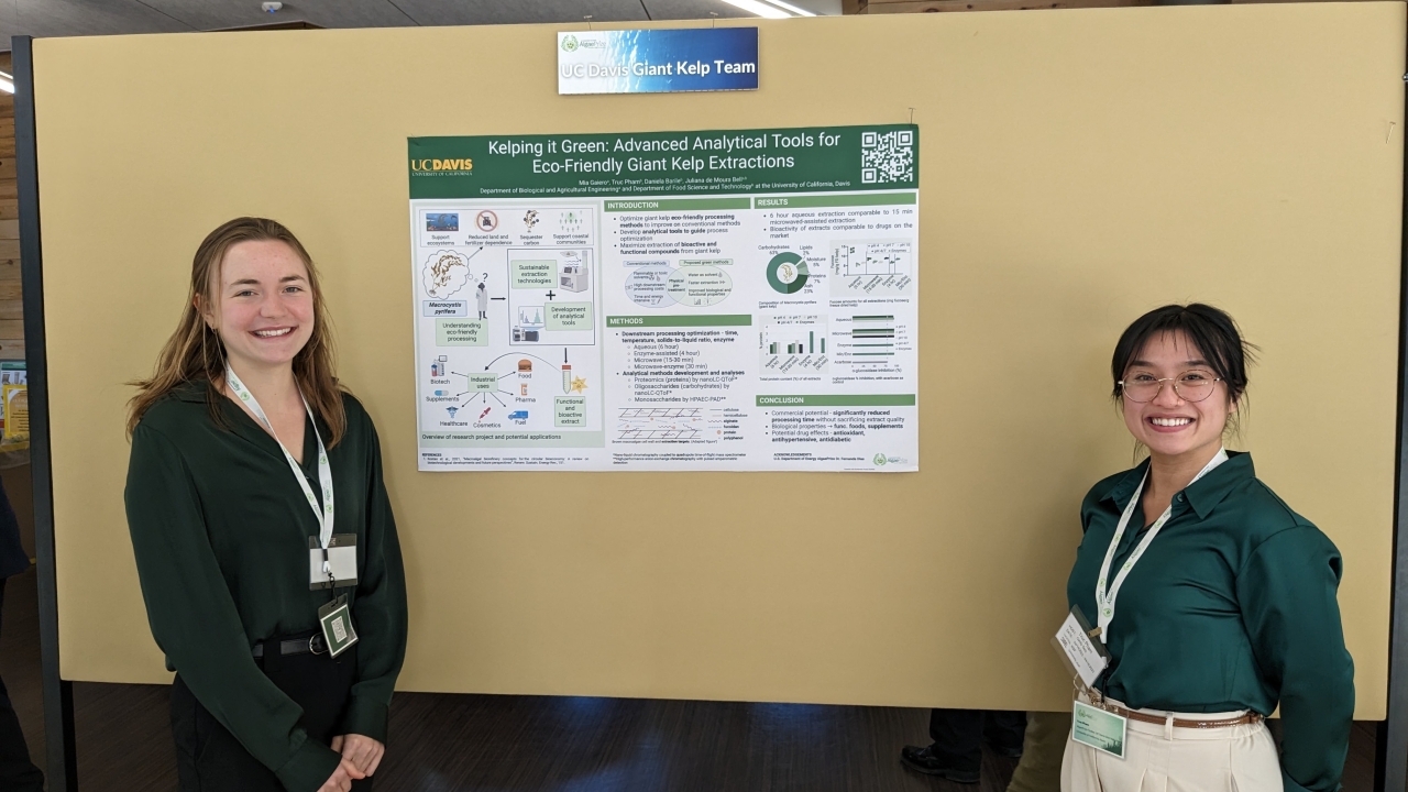 Two UC Davis students wear green button up shirts and lanyards while standing near a research poster