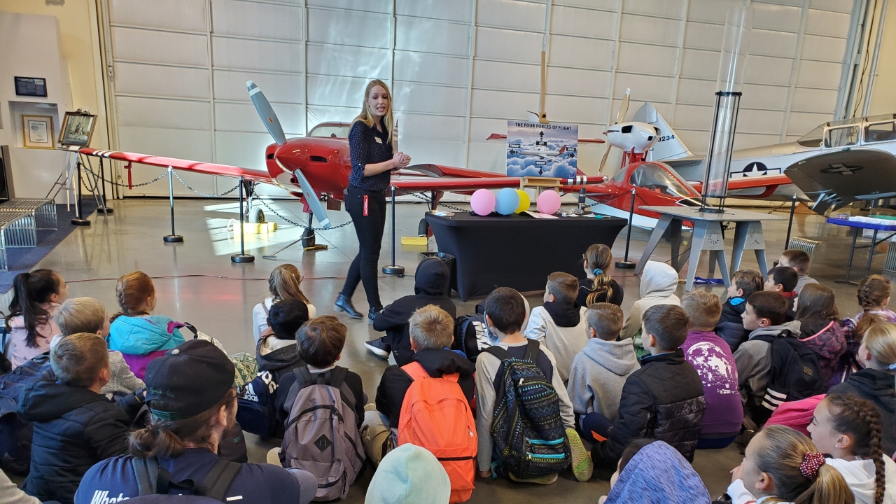 Students sitting on the ground while paying attention to a speaker near an airplane in a museum