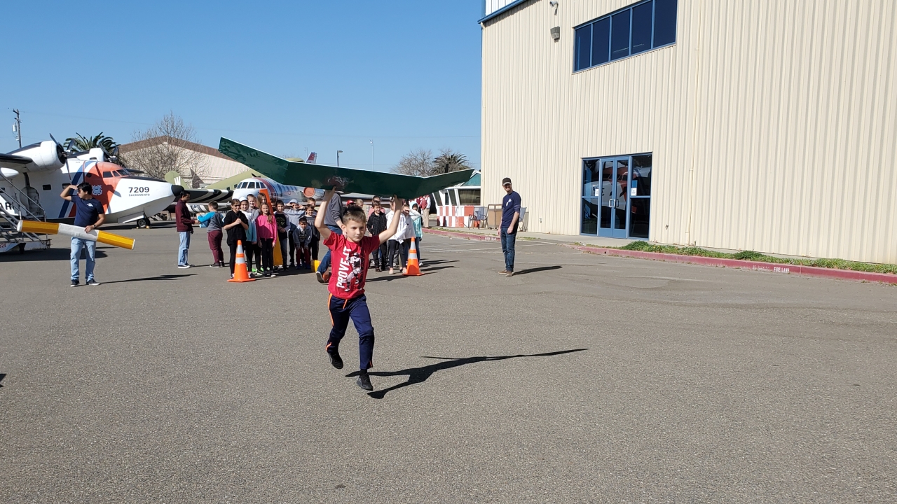 A young student holds a large green piece of aerospace material and runs away from a group of students behind cones