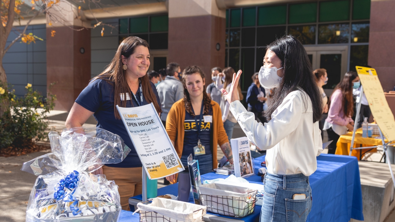 Two people stand behind tables with blue tablecloths and a gift basket on them and talk with another person, wearing a mask and waving