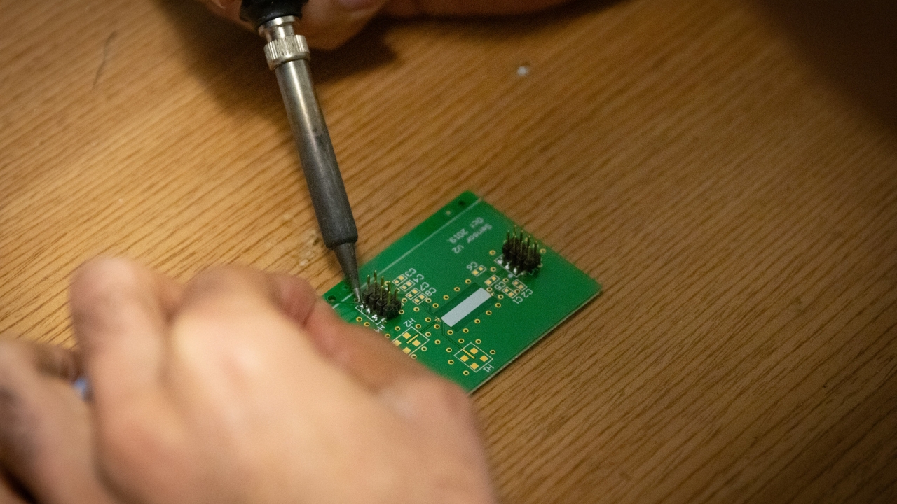 A green microchip sitting on a wooden table is worked on by a pair of hands holding a tool