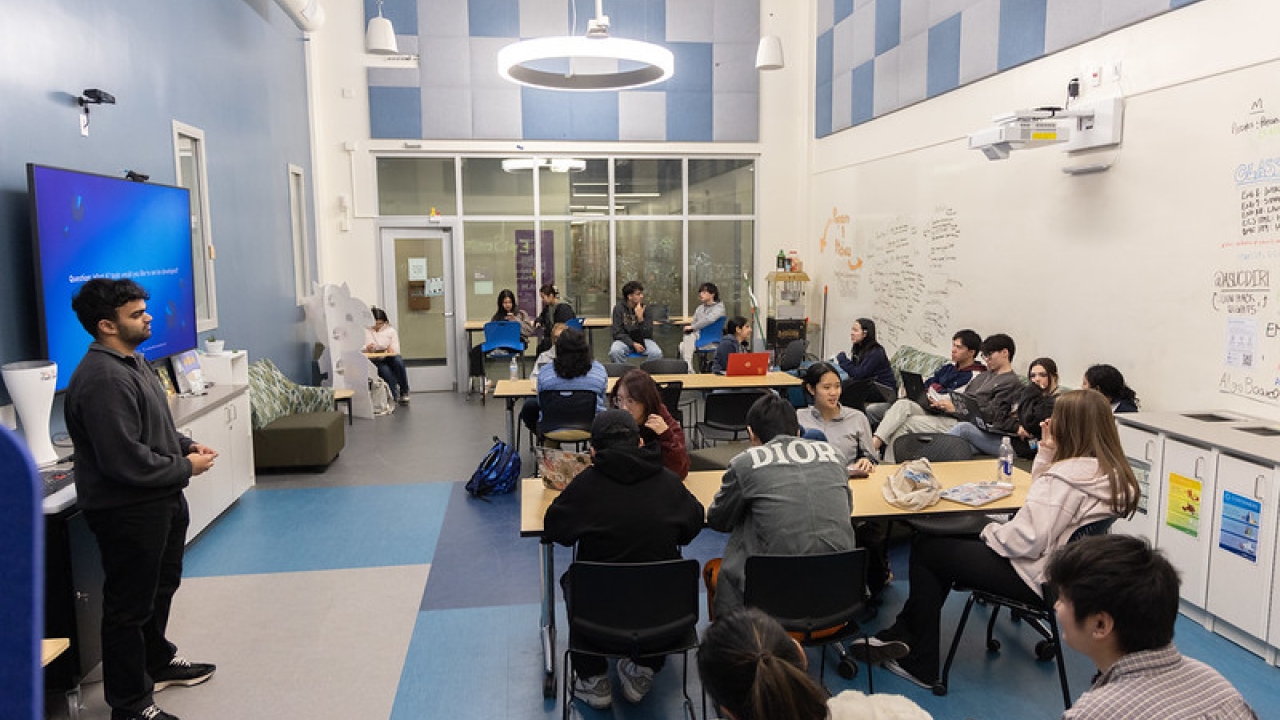 Students sit at tables while a student stands at the front of the class in front of a blue-screened monitor