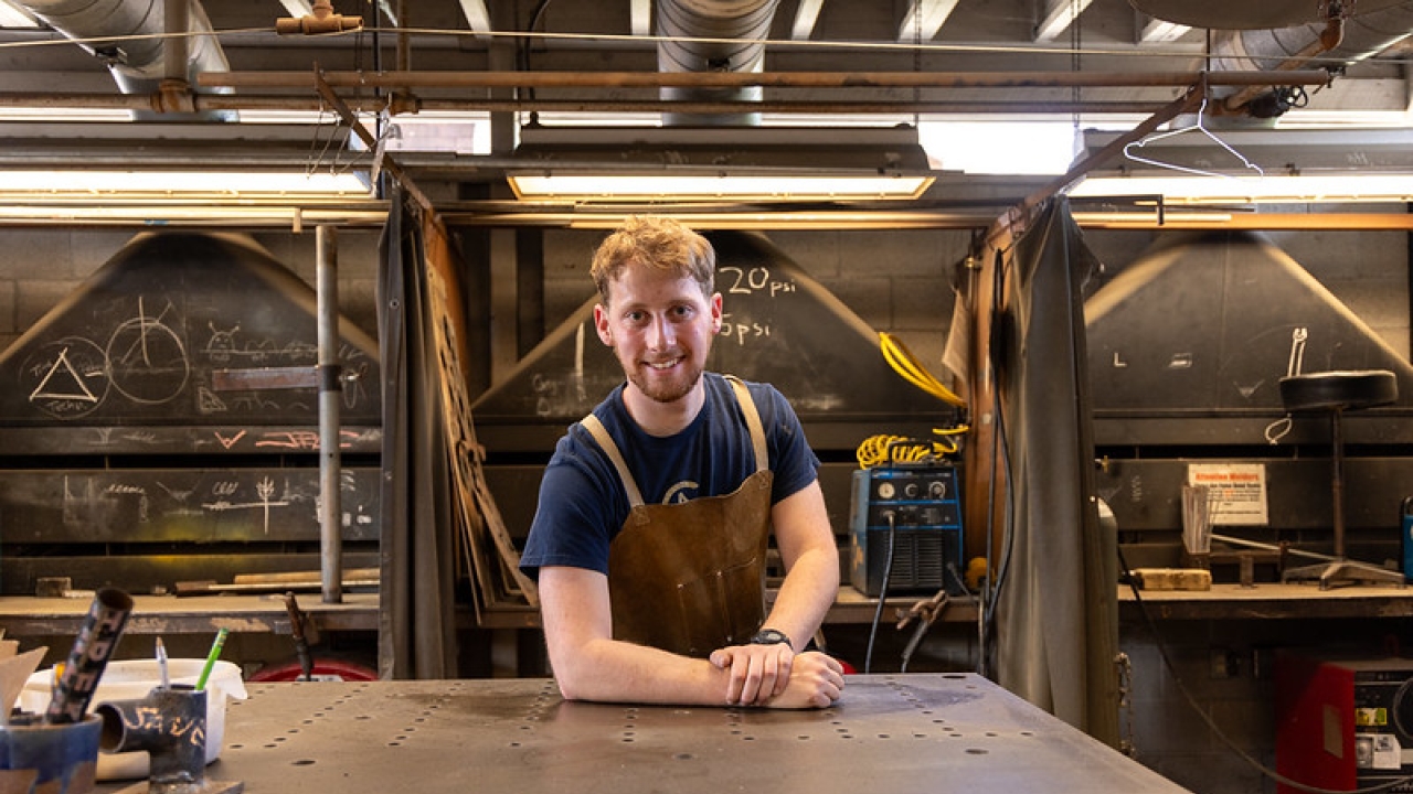 A young man with red hair wears an apron and stands in a welding studio