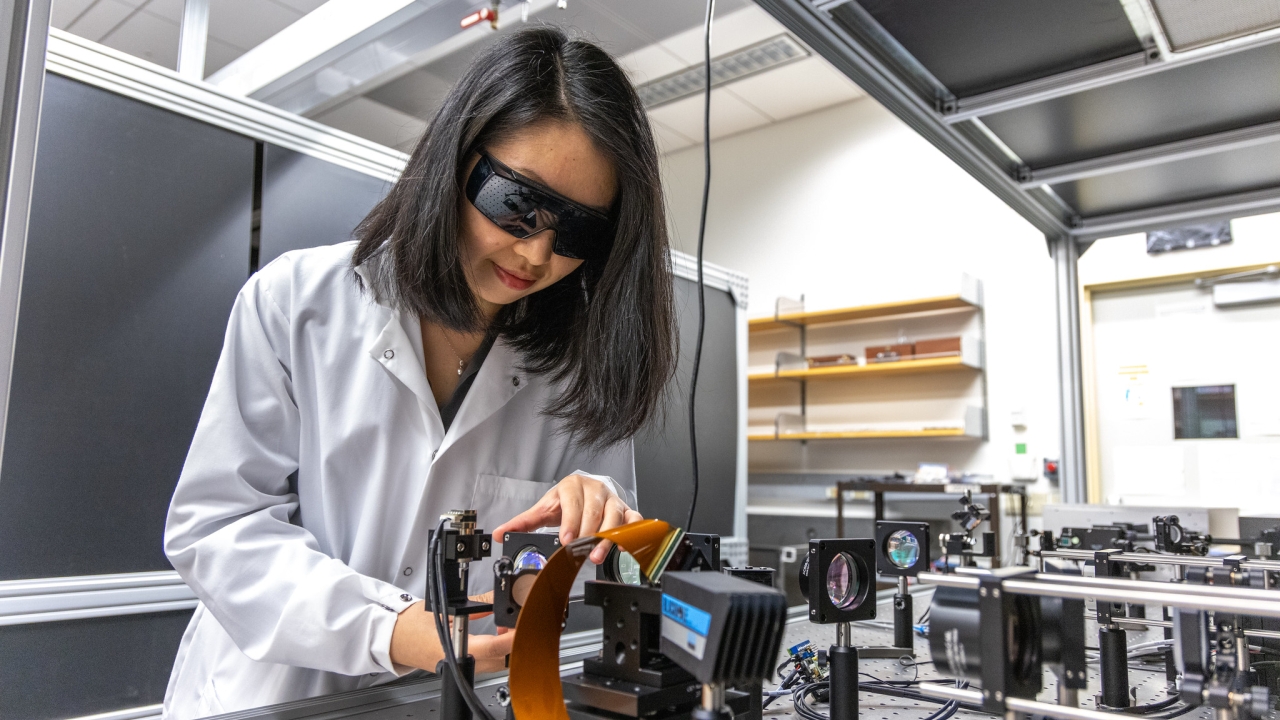 A woman in lab gear leans over a workbench 