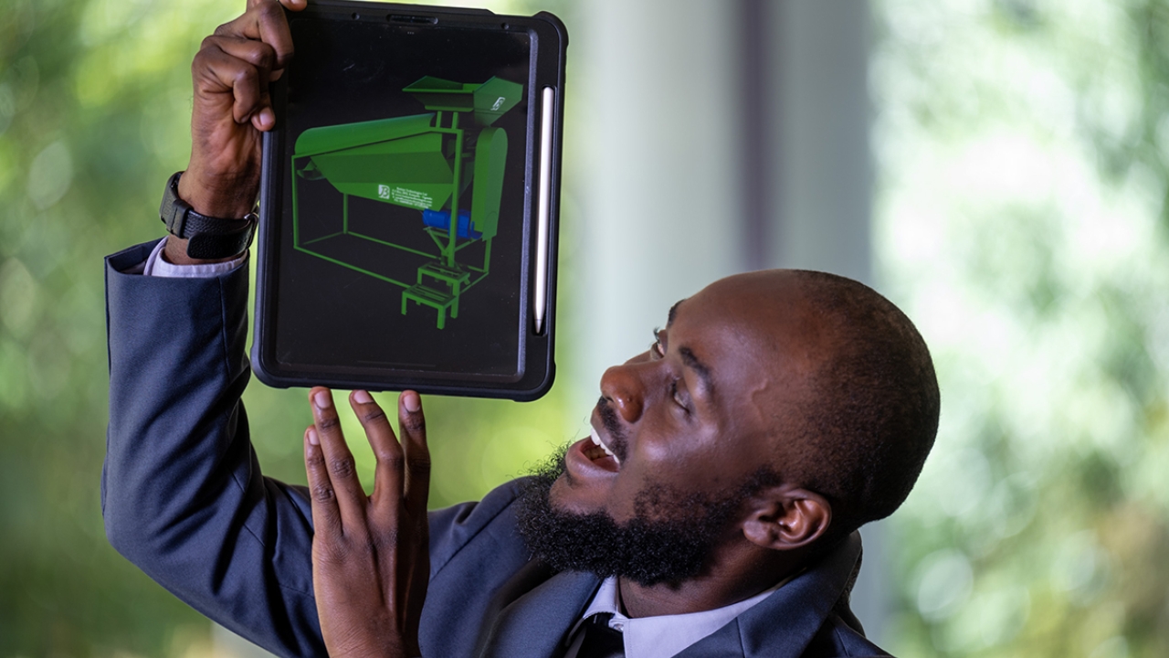 Ismael Mayanja wearing a suit holds up a tablet with a green model displayed