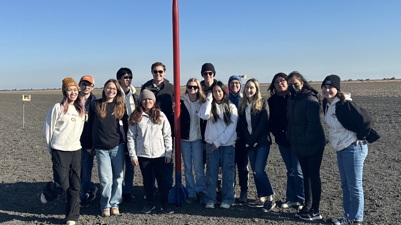 A group of students in an empty field standing around a tall, thin, red rocket