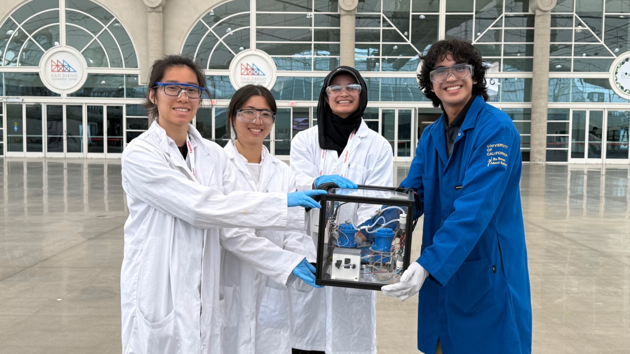 Four students in lab coats hold a cube structure in front of a building with windows