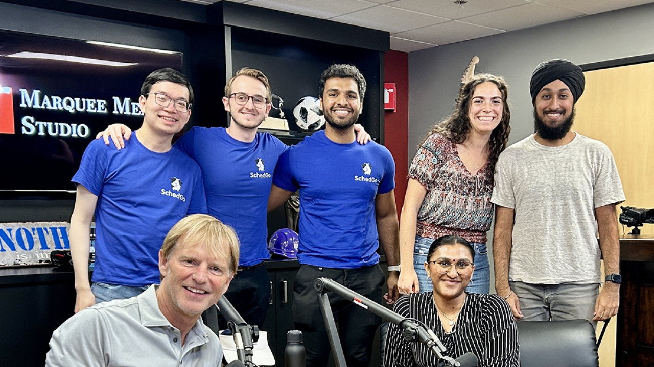 Mark Haney sits in a radio studio surrounded by several Square Solutions founders, students and entrepreneurs Henry Yu, Dillon Hill, Akshaj Raghavi, Sophie Bloyd, Harjn Bains and Deesha Patel. 
