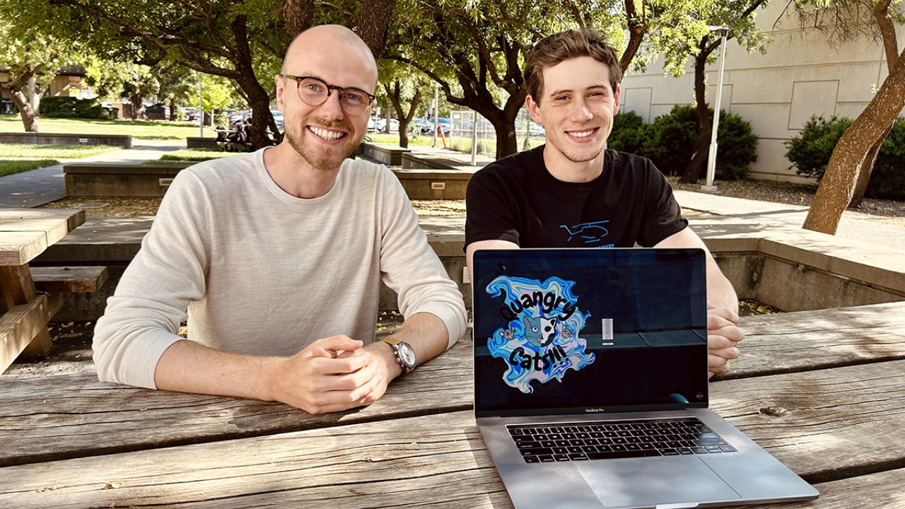 Quantum Computing at Davis members sit at an outdoor picnic table behind a laptop that has a black screen with blue colorful swirls and "Quangry Cats" written out