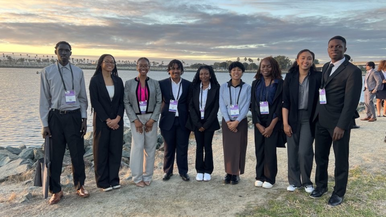 A group of Black students wearing nametags and lanyards stand on grass and sand