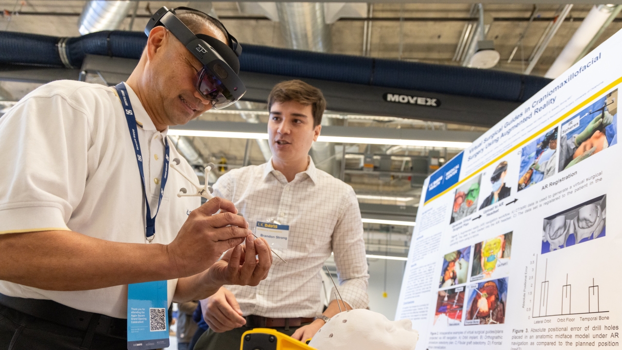 One person wears augmented reality goggles while another stands close by near a poster and interactive activity