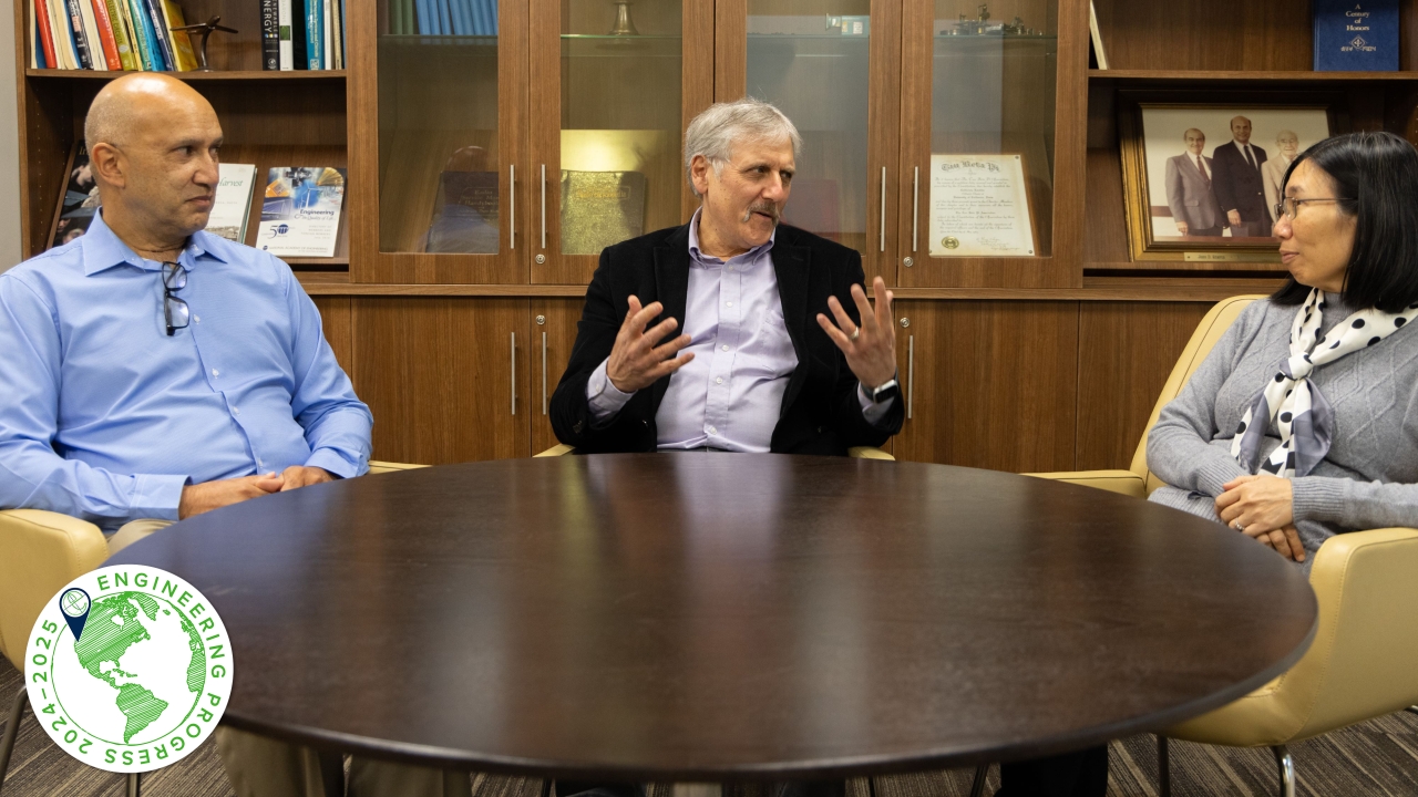Three individuals are seated at a round wooden table engaged in a discussion. Behind them, a wooden cabinet displays books, awards, and a framed photograph. The person in the center gestures with open hands, emphasizing a point, while the others listen attentively. In the lower-left corner, a circular logo with a globe design and the text 'Engineering Progress 2024-2025' is visible.