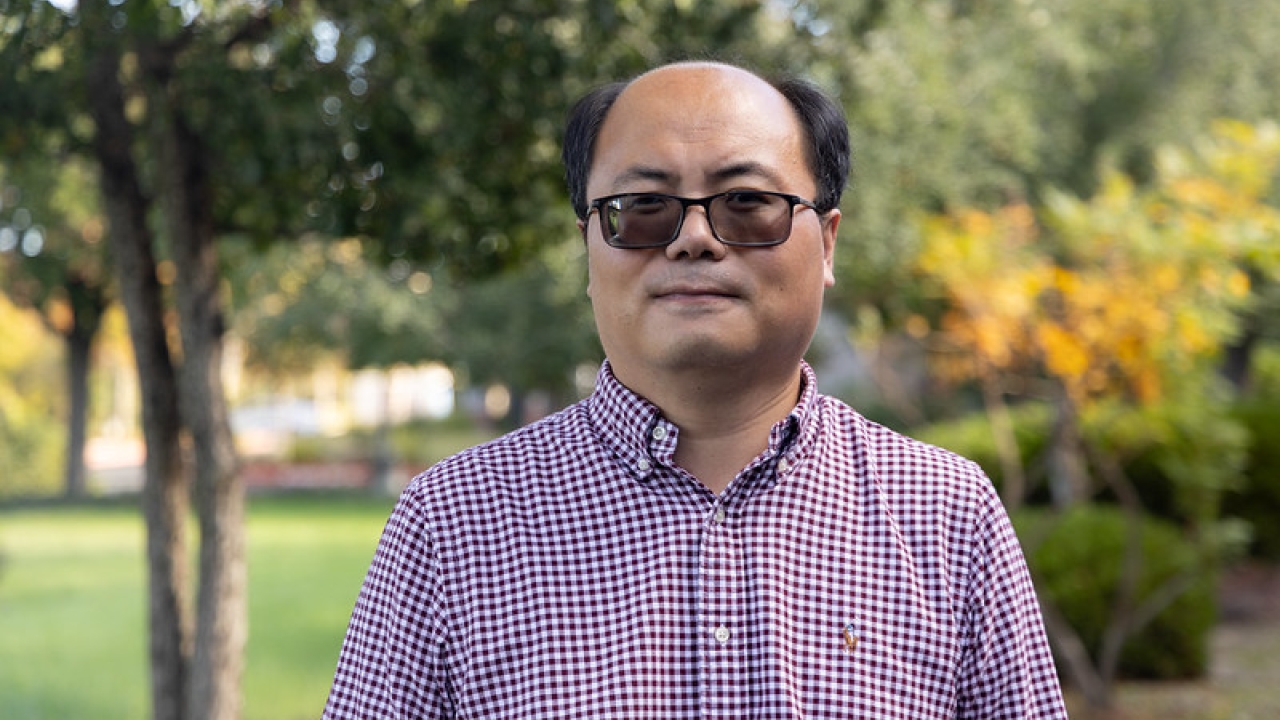 Zhaodan Kong smiles outdoors, wearing a checked shirt, surrounded by greenery.