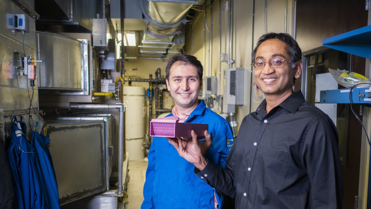 Two researchers hold a pink device standing in front of large tanks in a laboratory