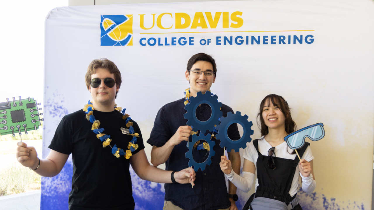 Three smiling young adults holding gears and circuit props in front of UC Davis College of Engineering banner