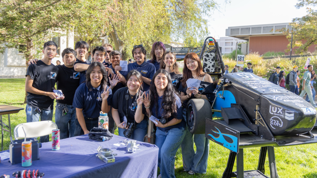 Group photo of smiling students around a blue electric car on a sunny campus lawn