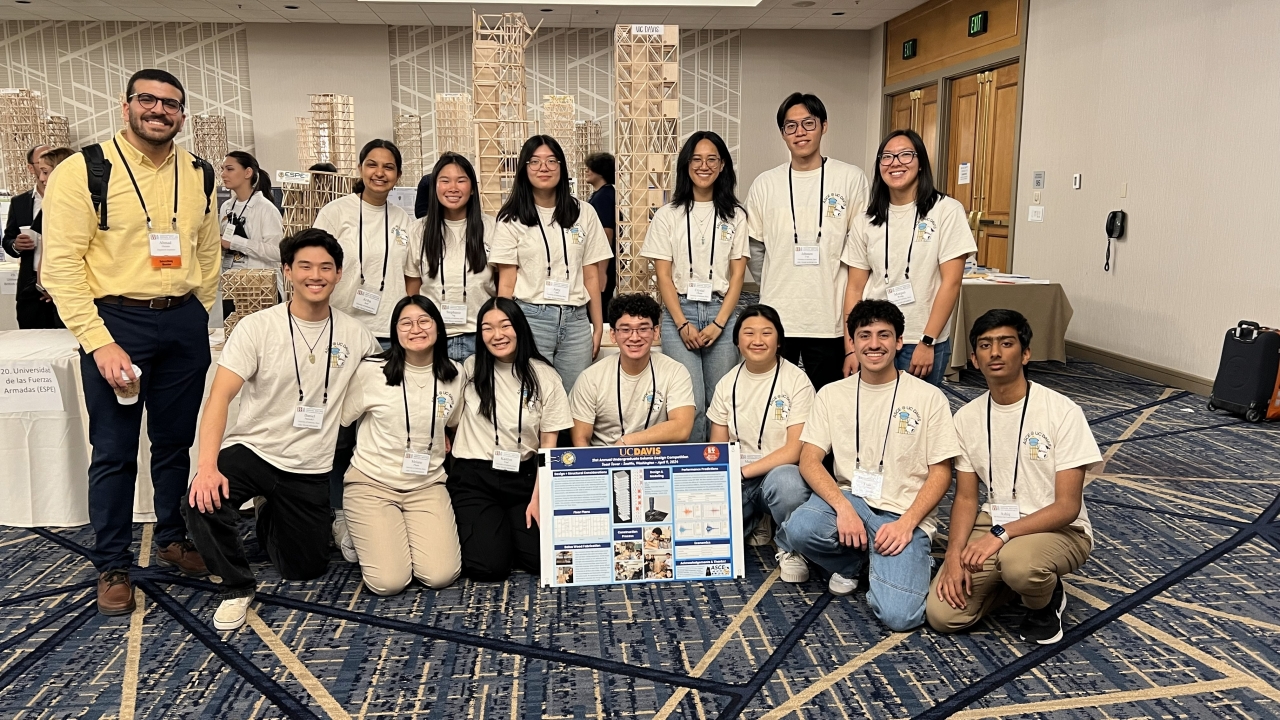 A group of students in matching shirts poses with a project display at a conference.