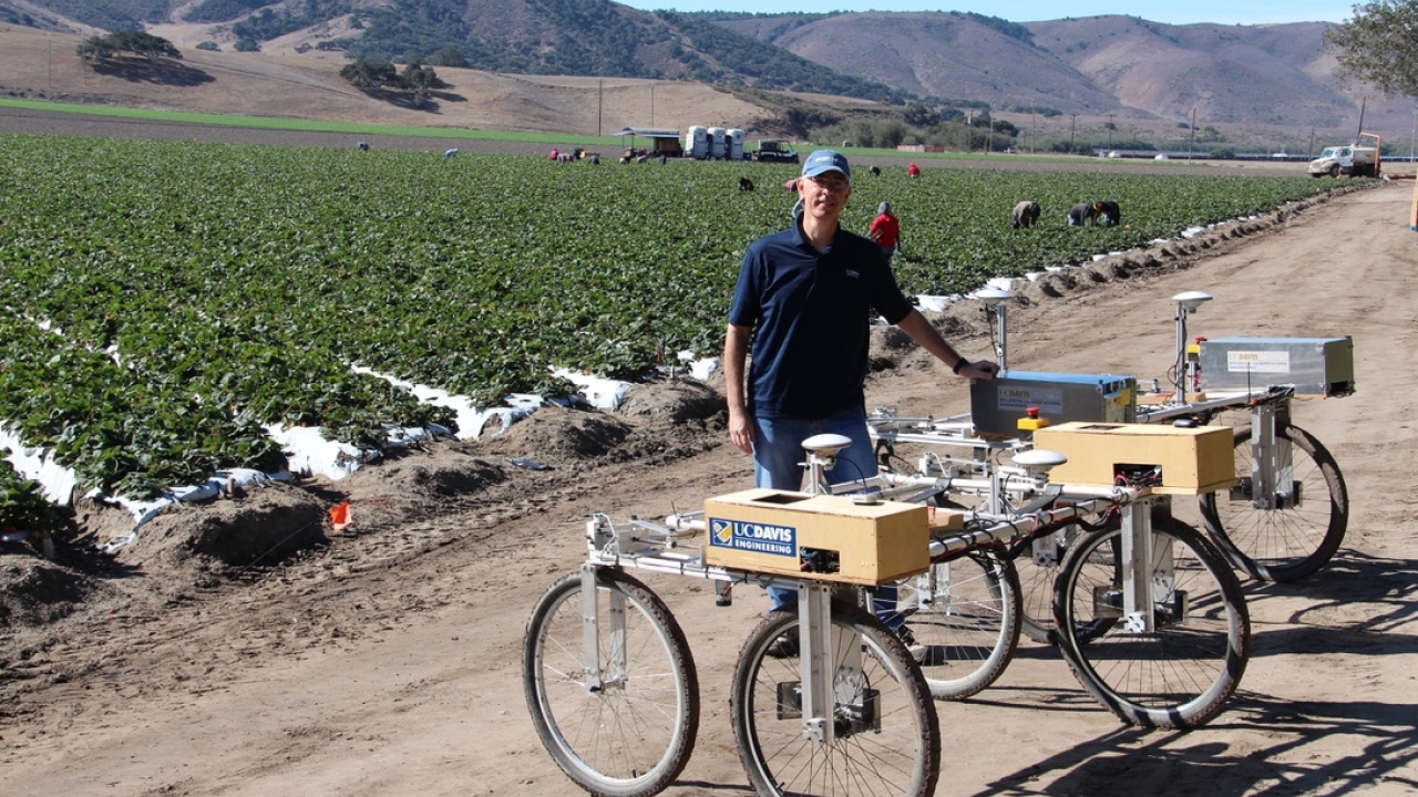 UC Davis engineer stands beside an agricultural robot in a strawberry field, with rows of plants and farmworkers visible in the background. The robot, labeled 'UC Davis Engineering,' has a metal frame with large wheels and mounted sensors used for agricultural research.