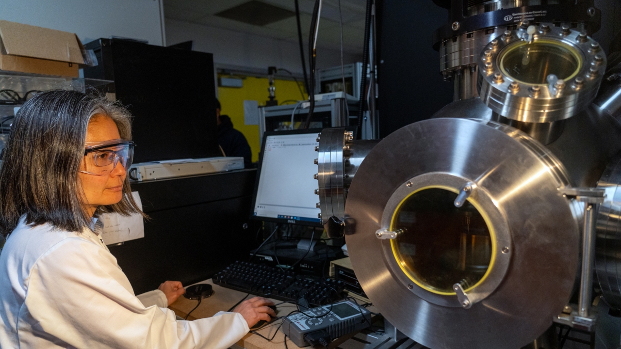 Yayoi Takamura, professor and chair of materials science and engineering wears safety goggles and a lab coat and stands near a machine in her lab