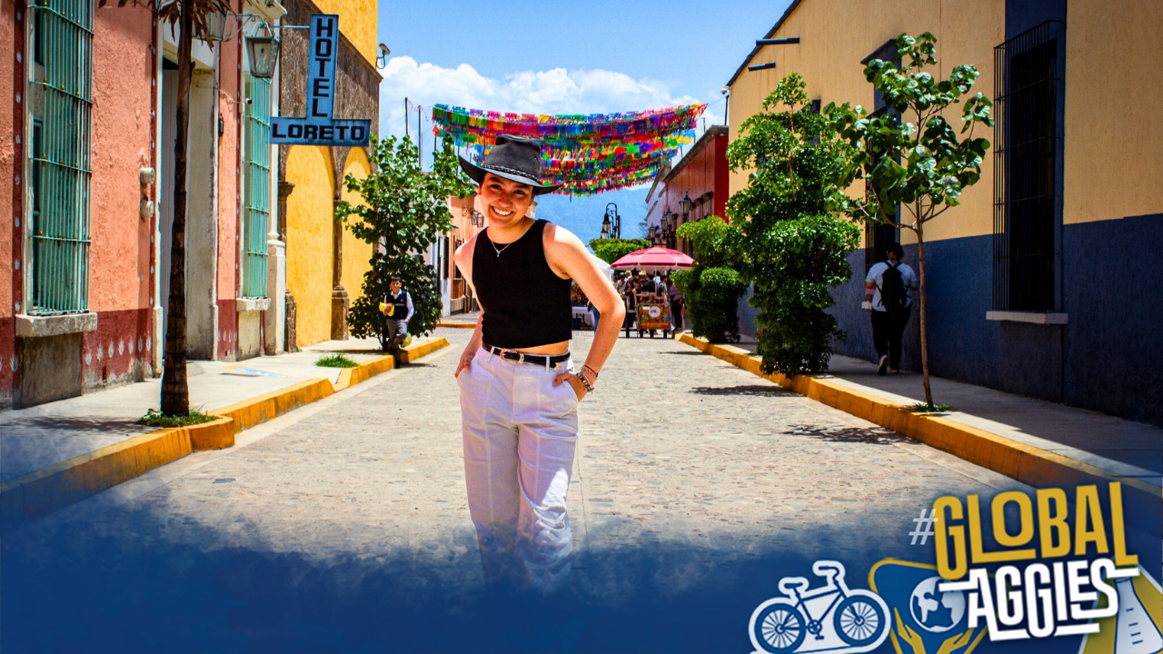 A smiling person in a stylish outfit walks down a colorful, sunny street with decorations above. Text in the bottom right corner says "Global Aggies".