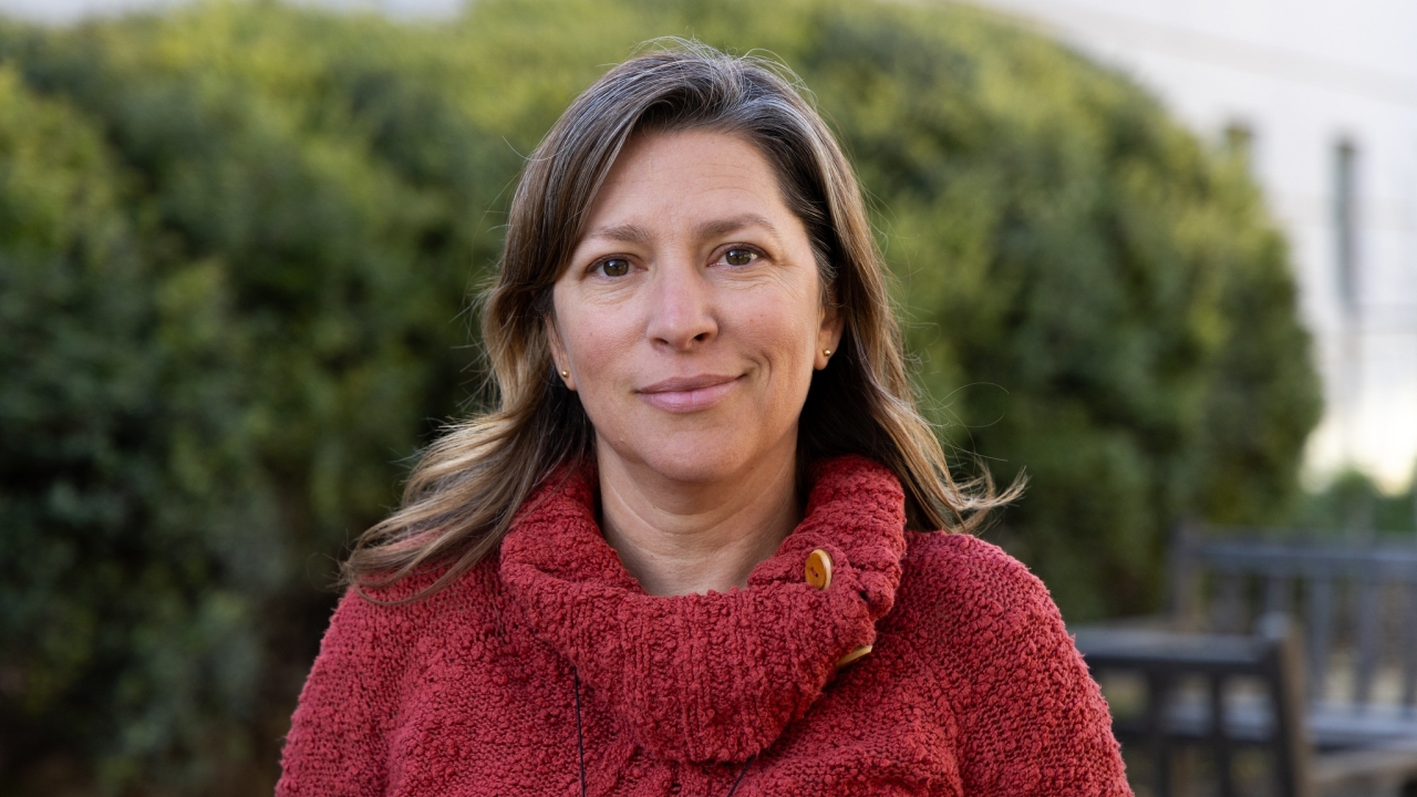 A woman with shoulder-length brown hair and light brown eyes stands outdoors in front of green shrubs and a blurred building. She wears a textured red sweater with a cowl neck and wooden buttons. She has a calm, confident expression with a slight smile and natural light highlights her face.