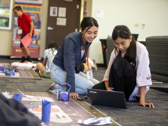 Two girls sitting on the ground with a laptop and materials