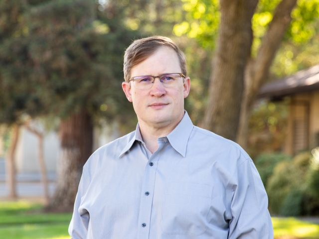 A man wearing glasses stands in front of a tree