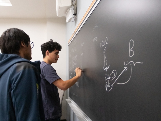 Two students stand by a blackboard, one student is writing in chalk