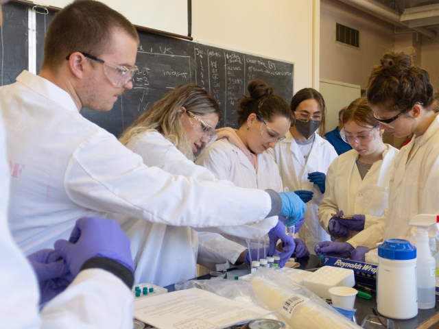 Group of students dressed in lab coats and PPE huddle around a table in a lab working on cocktail chemistry.