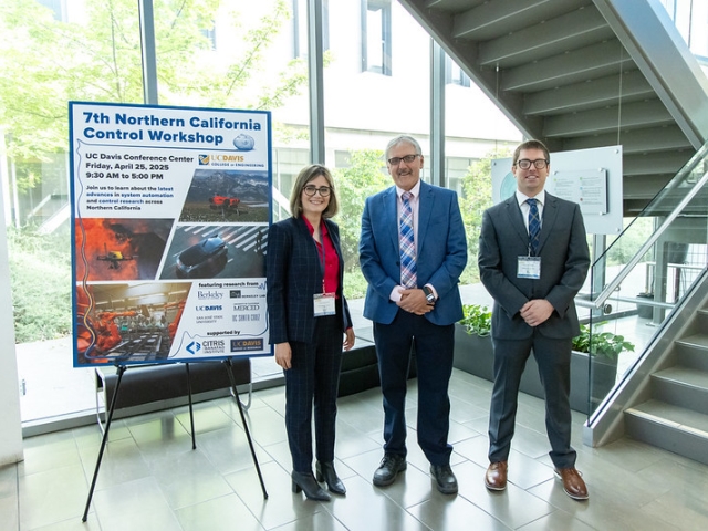 Three people stand indoors near a scientific poster and stairs