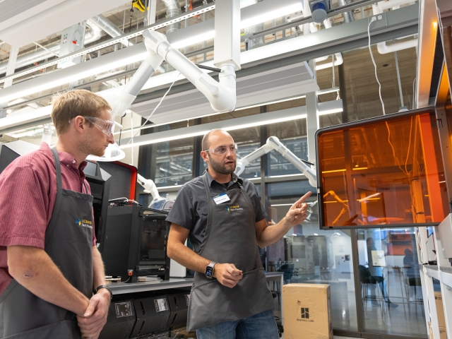 Two technicians in aprons discuss work in front of orange 3D printer